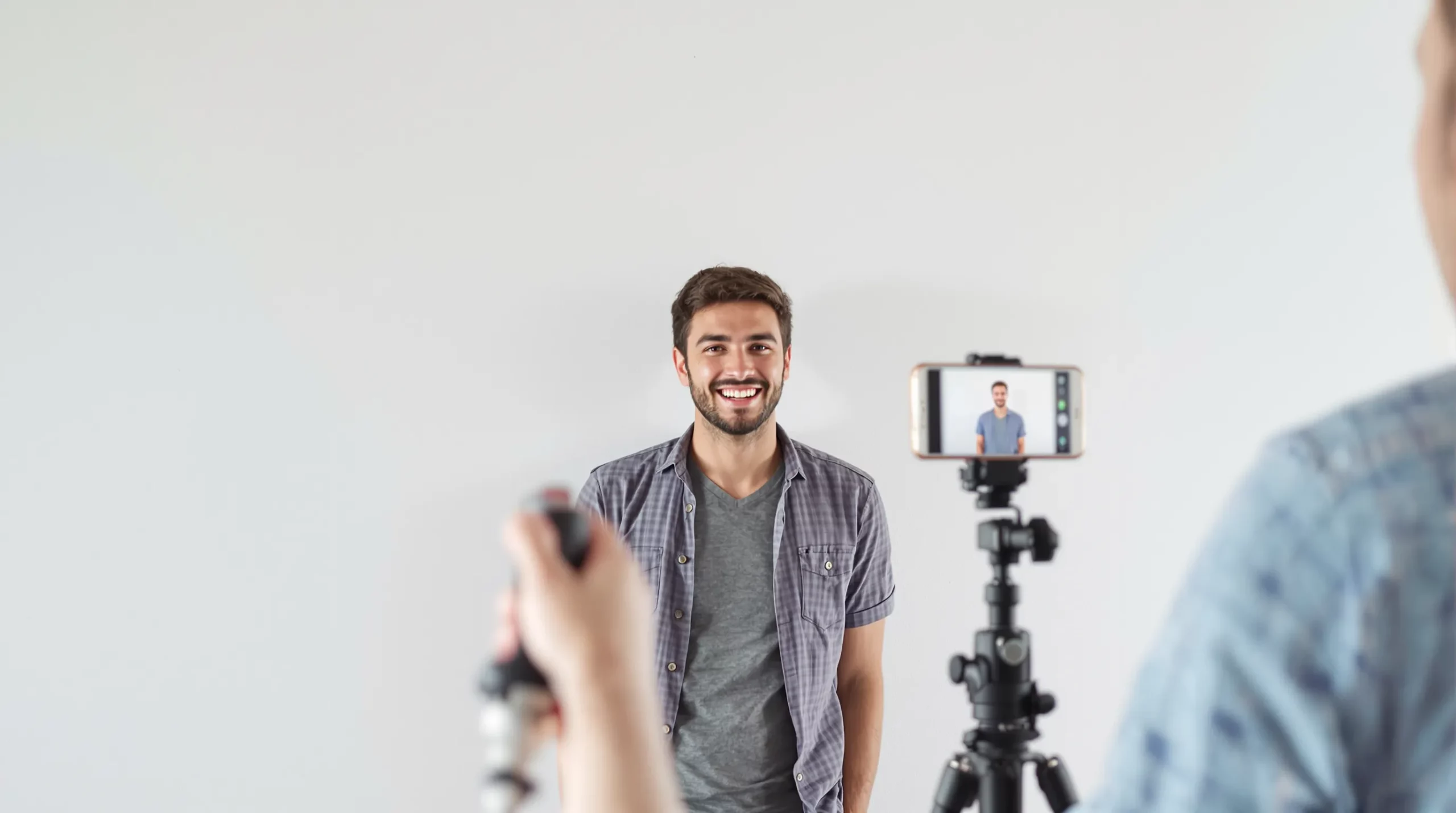 A traveler stands in front of a plain white wall with soft daylight from a nearby window, while a friend frames a head-and-shoulders photo using a smartphone on a tripod, ensuring even lighting and no shadows.