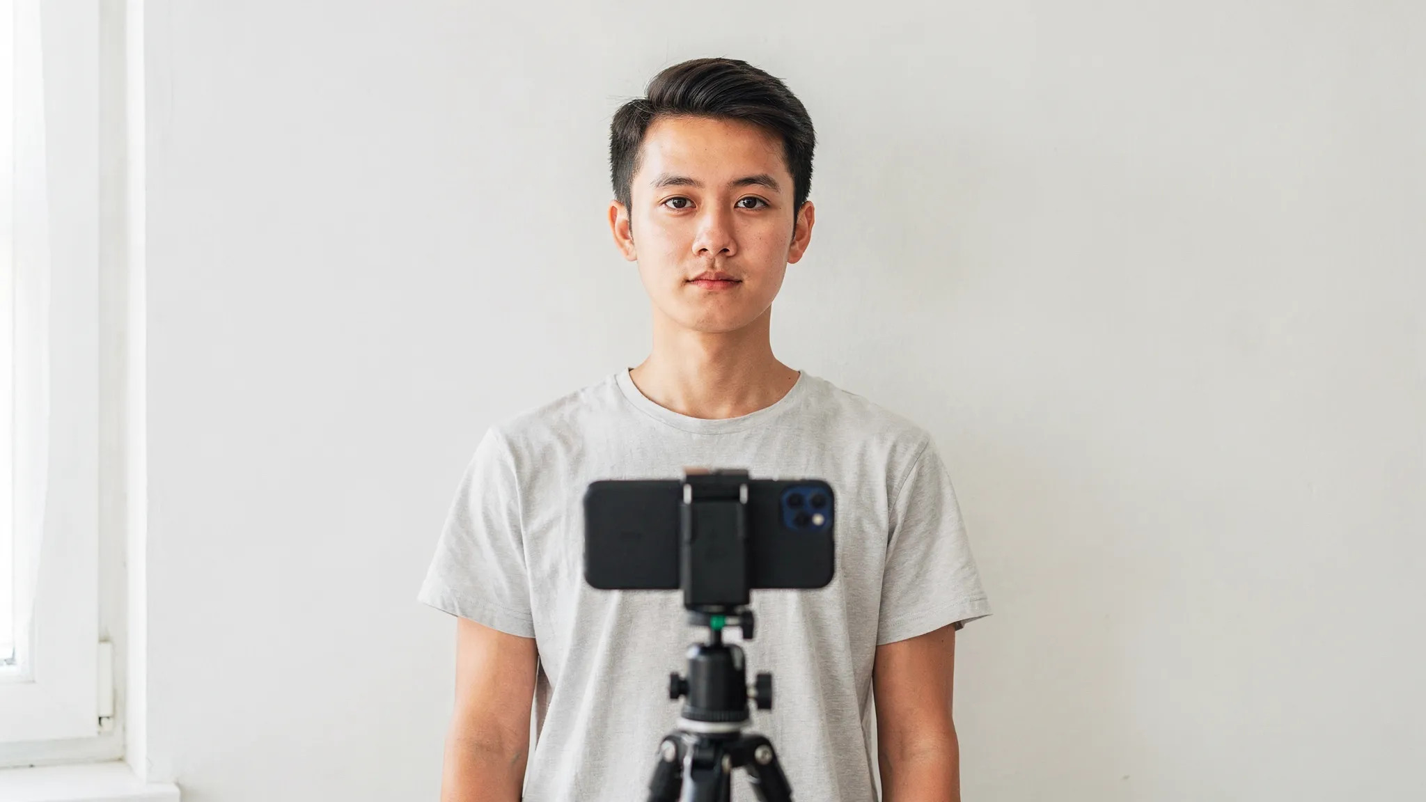 A simple at-home visa photo setup: a person standing 2–3 feet in front of a plain white wall, facing a bright window for even light, with a smartphone camera on a tripod at eye level. The background is clean with no decorations, and the person’s shoulders and head are centered in frame.