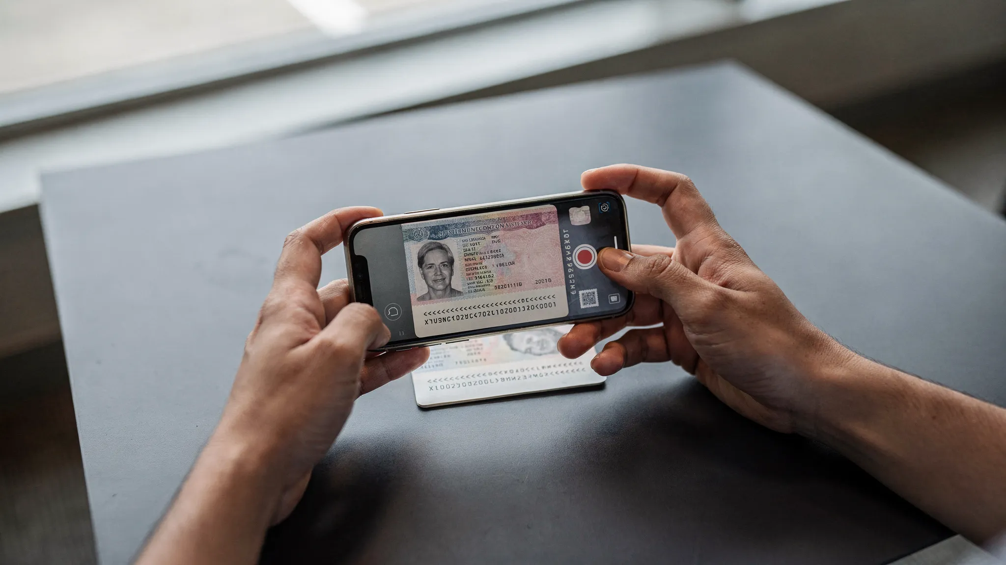 A traveler photographing a passport on a matte dark surface near a window, with the phone held parallel to the passport page and the MRZ in focus, showing soft indirect daylight and no flash glare.