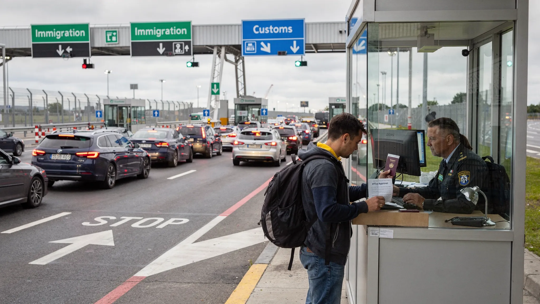 A busy land border checkpoint with cars and a pedestrian lane. A traveler holds a passport and a printed eVisa approval page while an officer checks documents in a small booth. Road signs indicate immigration and customs lanes.